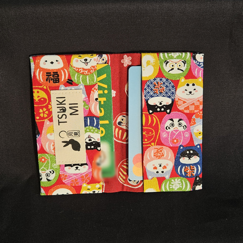 Porte-cartes Tsukimi darumas et shiba en motif dense sur fond rouge, ouvert avec deux cartes glissées dans les compartiments