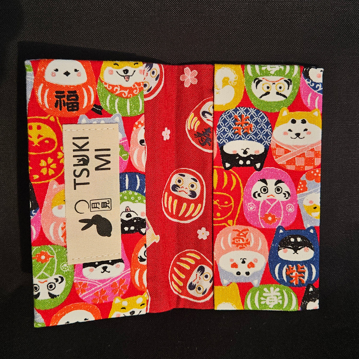Porte-cartes Tsukimi ouvert : extérieur petits darumas et shiba en motif dense, intérieur rouge foncé à darumas et fleurs de cerisier